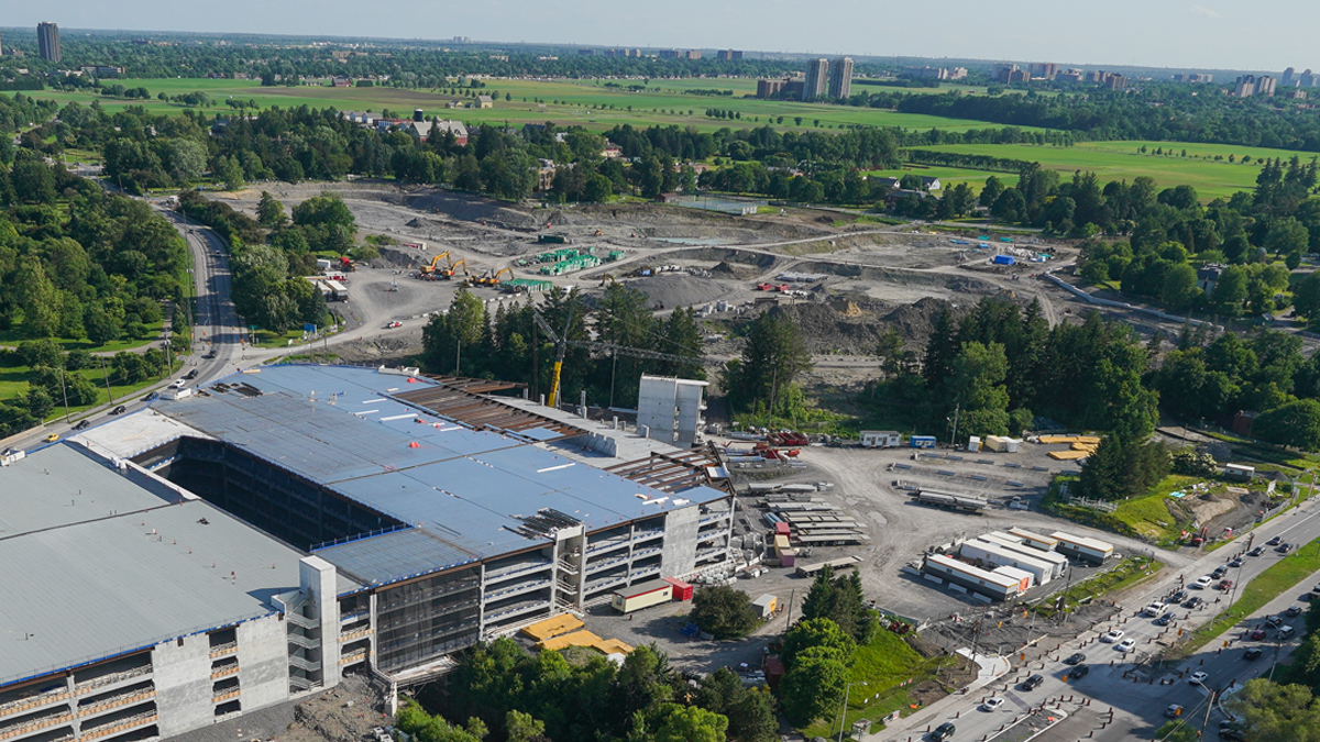 Alt tag: Vue aérienne du chantier de construction du nouveau campus de L’Hôpital d’Ottawa. Un stationnement à étages est en cours de construction en bas à gauche, et une vaste zone de terrain est en cours d’excavation en haut au centre. Le site est entouré d’arbres et de champs herbeux.