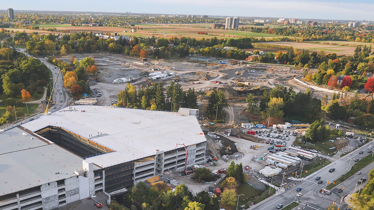 Alt tag: Vue aérienne du chantier de construction du nouveau campus de L’Hôpital d’Ottawa. Un stationnement à étages est en cours de construction en bas à gauche, et une vaste zone de terrain est en cours d’excavation en haut au centre. Le site est entouré d’arbres et de champs herbeux.