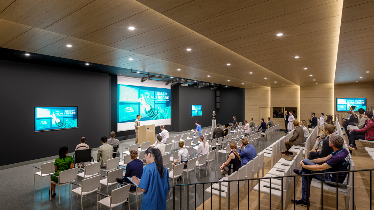 Concept artistique de l’intérieur d’un auditorium d’un nouvel hôpital. Un homme parle sur un podium, et trois grands écrans sont accrochés au mur derrière lui. Il y a plusieurs rangées de sièges et plusieurs personnes assises dans la grande salle.